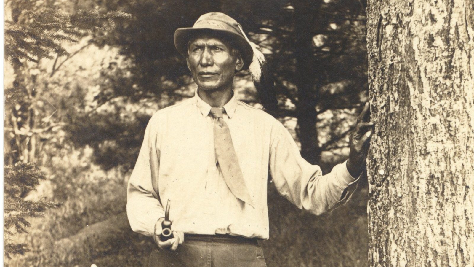 Charles Eastman with one hand on tree and other hand holding a smoking pipe