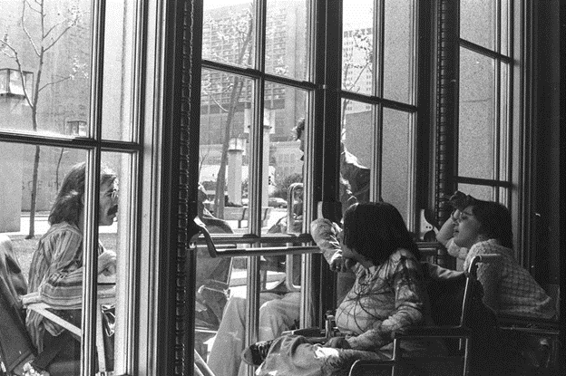 Black and white photograph of Mike Williams (left) on the outside of a glass door and Nancy D'Angelo (center) and Judy Heumann (right) on the inside of the door, all in wheelchairs, communicating with each other during 1977 sit-in protests.