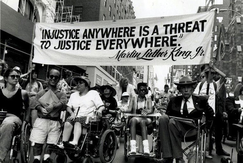 Black and white photograph of the 1993 Madison Square Disability Independence Parade. Judy Heumann and others march in their wheelchairs in front of a banner that reads 'Injustice anywhere is a threat to justice everywhere.' - Martin Luther King, Jr.