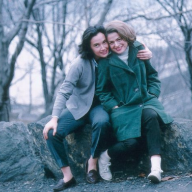 Thea Spyer (left) sitting with her arm around Edie Windsor (right) outside on some rocks with bare trees in the background.
