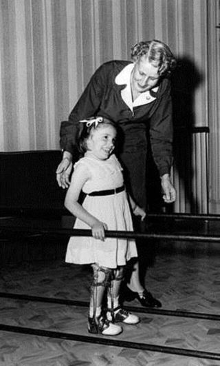 Black and white photograph of Judy Heumann as a young child using leg braces and a railing with the help of her mother, who is standing behind her.