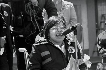 Black and white photograph of Judy Heumann wearing a Sign 504 button, speaking into a microphone.