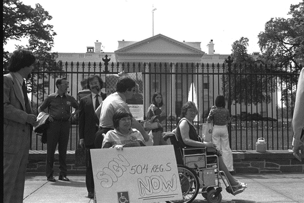 Black and white photo of protesters in front of the White House. Judy Heumann, center, sits in her wheelchair holding a sign that reads 'Sign 504 regs NOW.' alongside activist Kitty Cone.