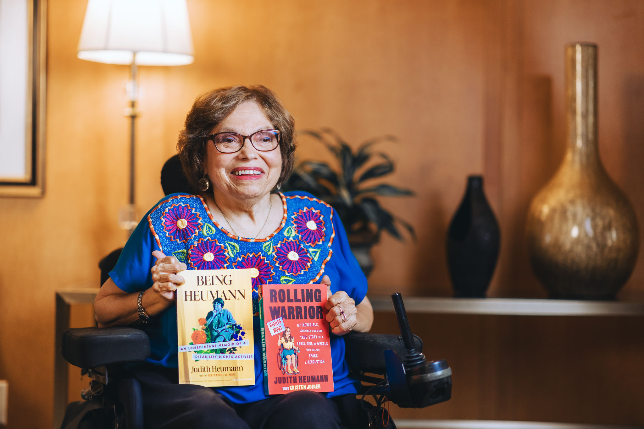 Color photograph of Judy Heumann in her wheelchair holding up copies of her two memoirs, 