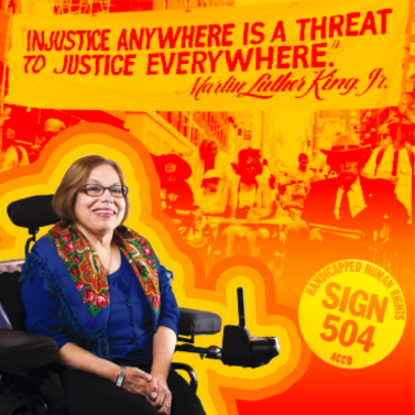 Photo of Judy Heumann in front of a sign reading Injustice anywhere is a threat to justice everywhere.