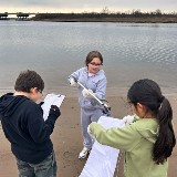 Three students at the beach stand in front of the water on the sand. The first student (left) is holding a clipboard and pencil, while the second student (center) places litter into a trash bag held by the third student (right), using a trash picker.