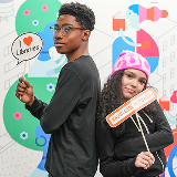 A teen boy (left) and girl (right) pose together in front of a colorful background featuring an illustrated figure reading a book, holding up signs that read 'I Love Libraries,' and 'Knowledge is Power,' respectively.