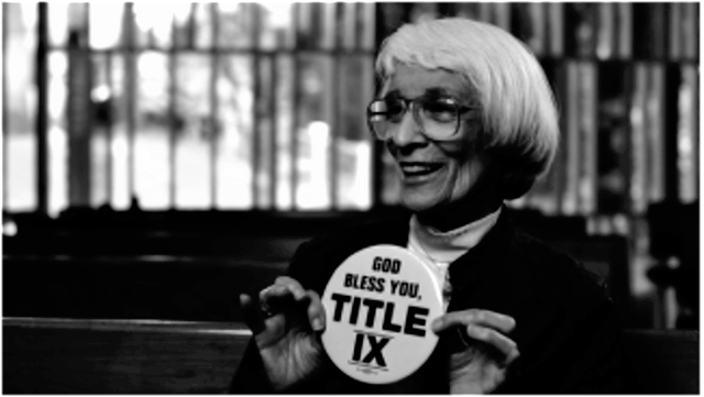 B&W photo of Bernice Sandler smiling and holding up a round badge with text reading "God bless you, Title IX"