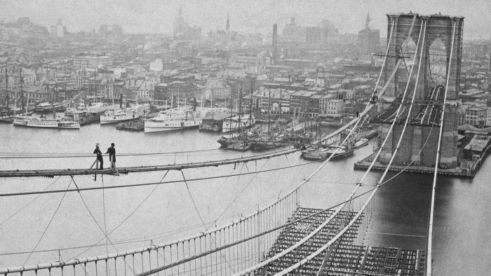 Black and white photo of the Brooklyn Bridge as it was under construction during the 1880's. Two men can be seen balancing on top of a row of suspension cabling.