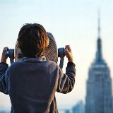 Behind-the-back view of a young child looking through a tower viewer to see the Empire State Building in the background.