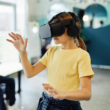 Close-up of a young girl wearing a VR headset; she has her hand in the air as if reaching out to touch something/someone.