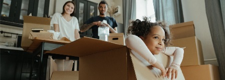 Young girl sitting inside an empty moving box in new home with family in background.