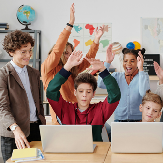 Two students sitting in front of laptops while students behind them cheer them on, and an adult standing on the left side.