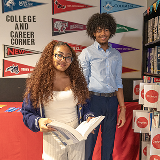 Two students in a library in front of a sign that reads 'College and Career Corner.' The student on the left is holding an open book.