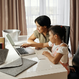 A mother and daughter sitting together at a table with a laptop. The daughter is pointing at the laptop screen.