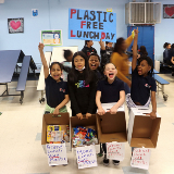 Five students at P.S. 188 The Island School in Manhattan hold up a sign with Plastic Free Lunch Day written on it, and display the plastic waste they have collected from their cafeteria, which is sorted into four cardboard boxes.