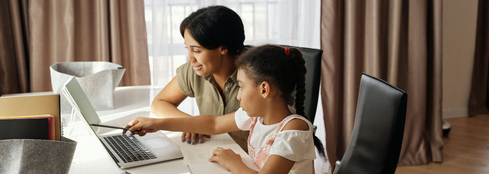 A mother and daughter sitting together at a table with a laptop. The daughter is pointing at the laptop screen.