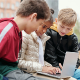 Three young kids looking at a laptop together, with the child in the middle in charge of typing.