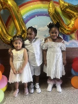 Three rising kindergarteners, a boy standing in between two girls, standing in front of a rainbow and a gold "2024" balloon set above.