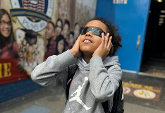 Student standing outside school building wearing eclipse glasses to look at the sun.