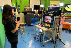 A student stands in from of a green screen to record an interview. She is facing a teleprompter, and another screen showing her video feed.