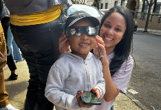 A young, smiling student wearing eclipse glasses, a grey sweatshirt, and a black hat poses for the camera with a school staff member.