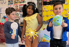 Three students in a classroom - from left to right, they are holding puppets representing the moon, sun, and earth for an eclipse activity.
