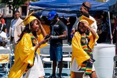 Side view of two young women walking, wearing yellow graduation caps and gowns adorned with black sashes featuring red, green, and yellow accents.