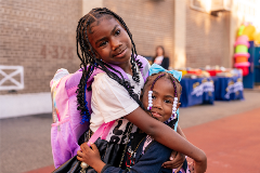 Two girls hugging one another; one on left is taller and older