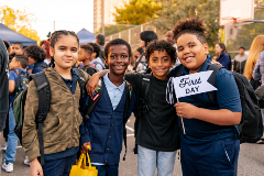 Four elementary-school aged students posing next to each other, with one holding a small flag that reads, "First Day."