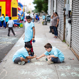 Two elementary school-aged students sitting down on sidewalk playing tic-tac-toe with each other with chalk while another student looks on.