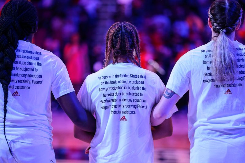 The backs of three women's basketball players standing arm in arm before tipoff wearing t-shirts containing text from the Title IX legislation
